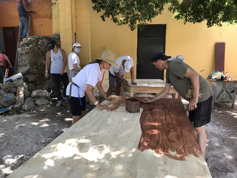 A man and a woman stain a large table while other people work in the background in the shade on a sunny day.