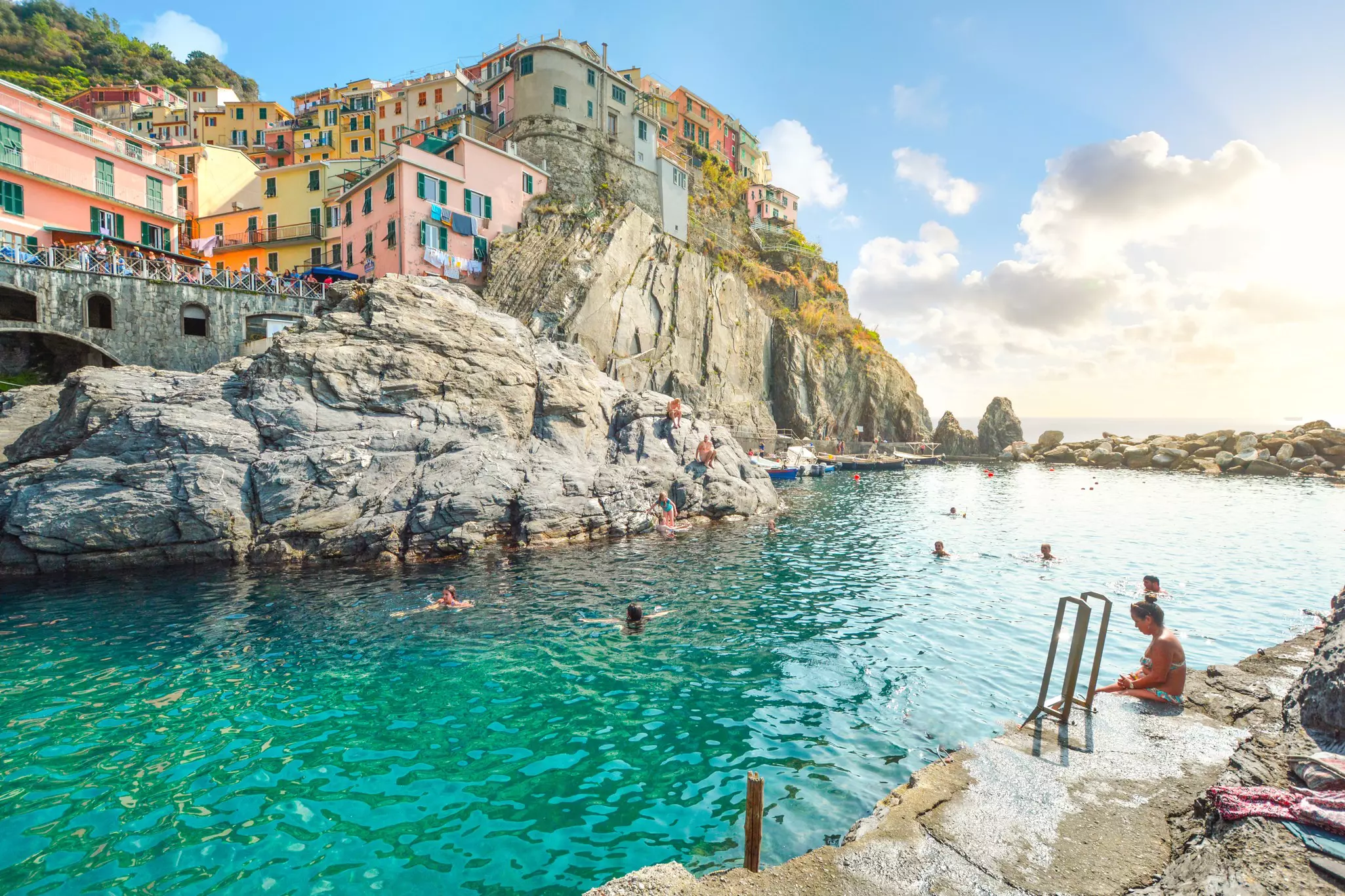 People swimming in a sheltered cove at the foot of a cliff stacked with pastel-colored buildings.
