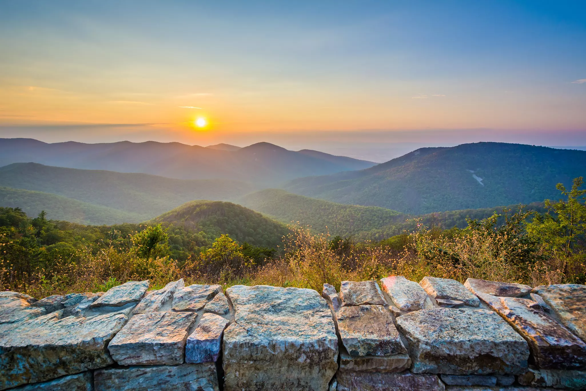 The silhouettes of mountains with a setting sun; a rock wall is in the foreground.