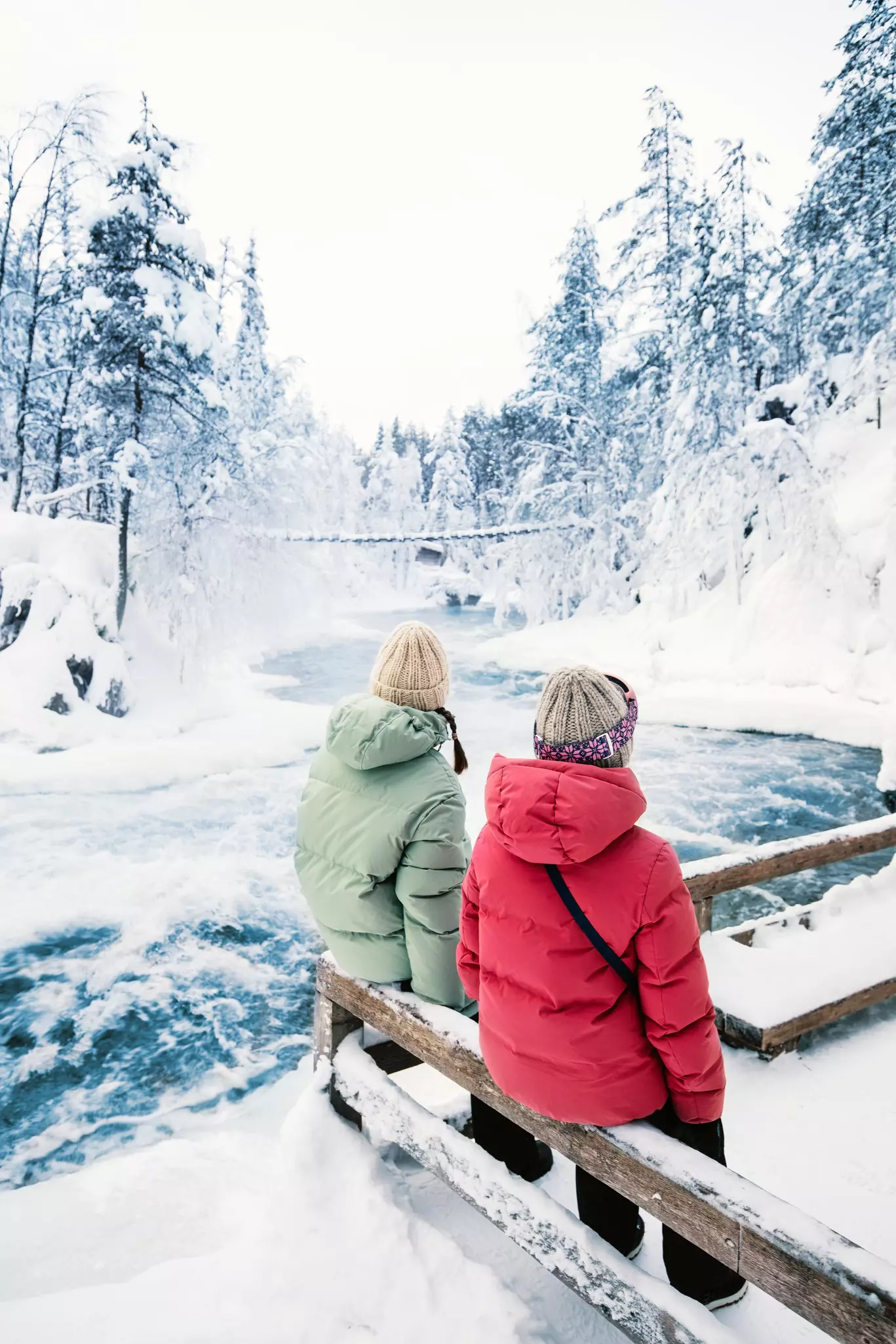 Two women on a snow covered wooden rail look out at a snowy wilderness scene with a blue frothy river flowing through.