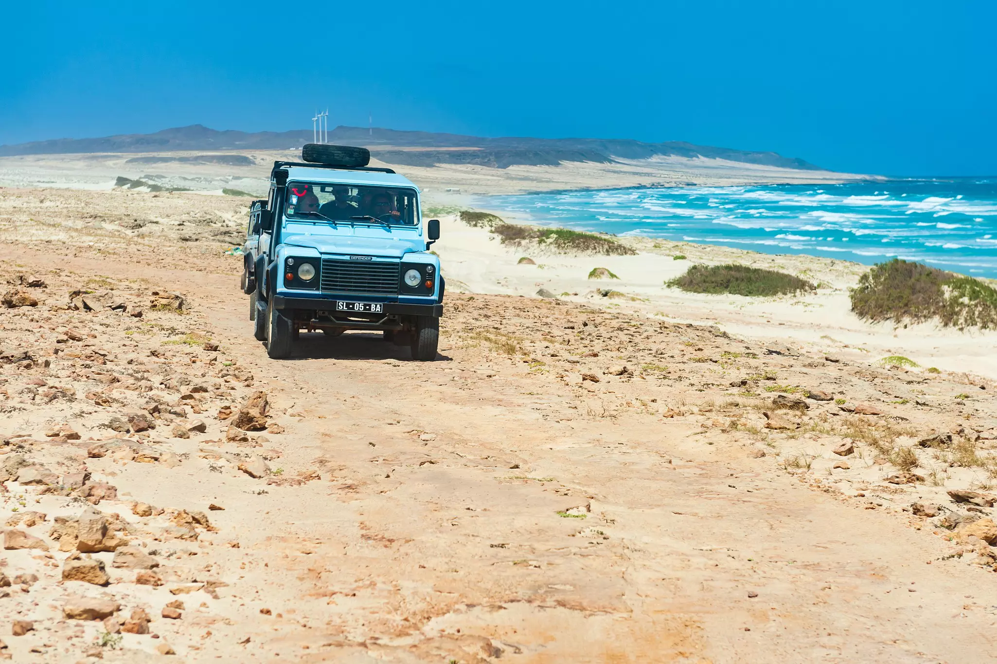 A four-wheel drive vehicle drives along a desert path next to the sea with waves crashing on the shore.