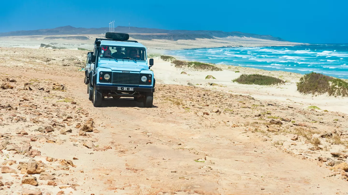 A four-wheel drive vehicle drives along a desert path next to the sea with waves crashing on the shore.