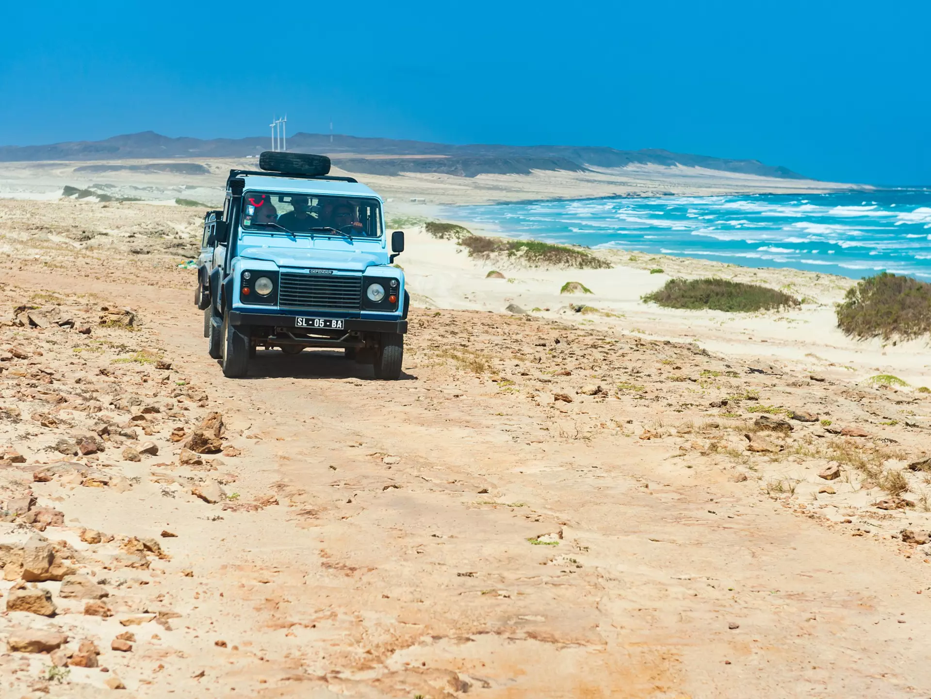 A four-wheel drive vehicle drives along a desert path next to the sea with waves crashing on the shore.