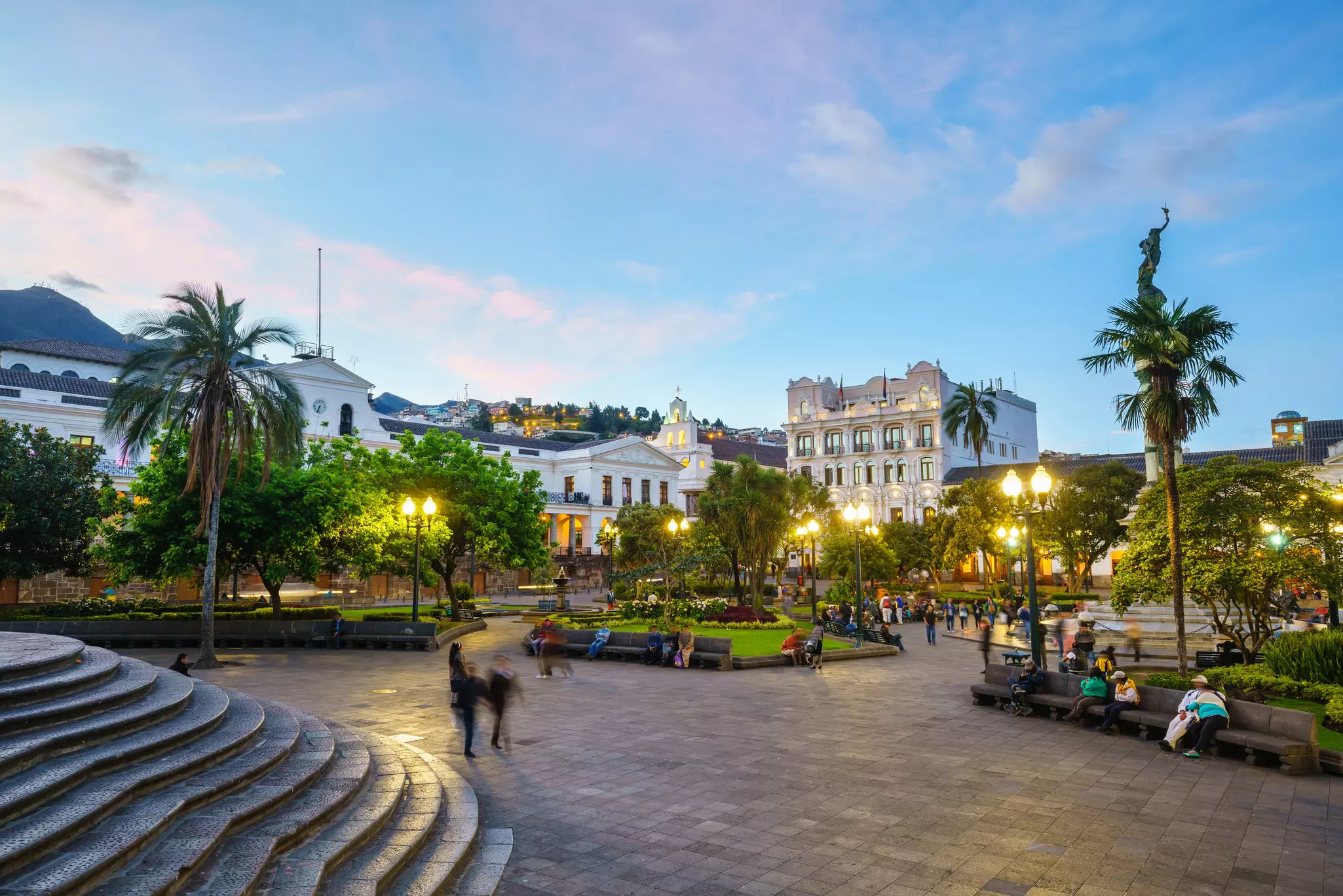 People walk and sit at dusk in a city square surrounded by historic buildings.