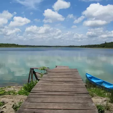Weathered wooden dock leading into a turquoise lake reflecting white clouds, with reeds and a low green treeline