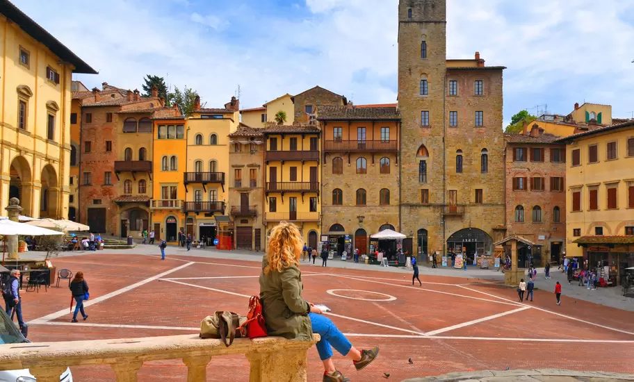 Woman sitting on a stone handrail facing towards a city square dominated by a tower