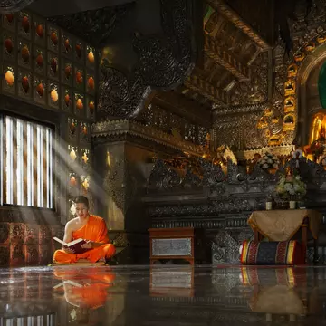 Young novice Buddhist monk, sitting cross-legged, reading inside a Chiang Mai monastery