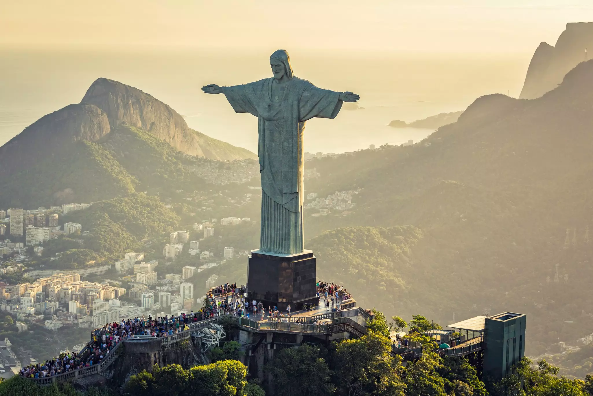 A monumental statue of Jesus Christ with arms outstretched at the top of a mountain. A crowd of people surrounds the statue's base; and a city is visible in the valley below.