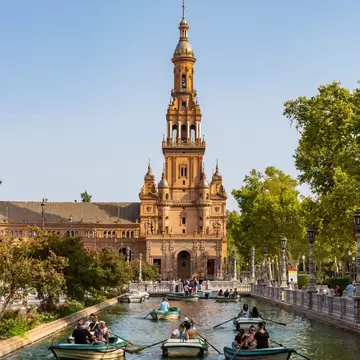 People rowing small boats along a canal toward an ornate brick tower, with leafy trees and balustrades on either side.