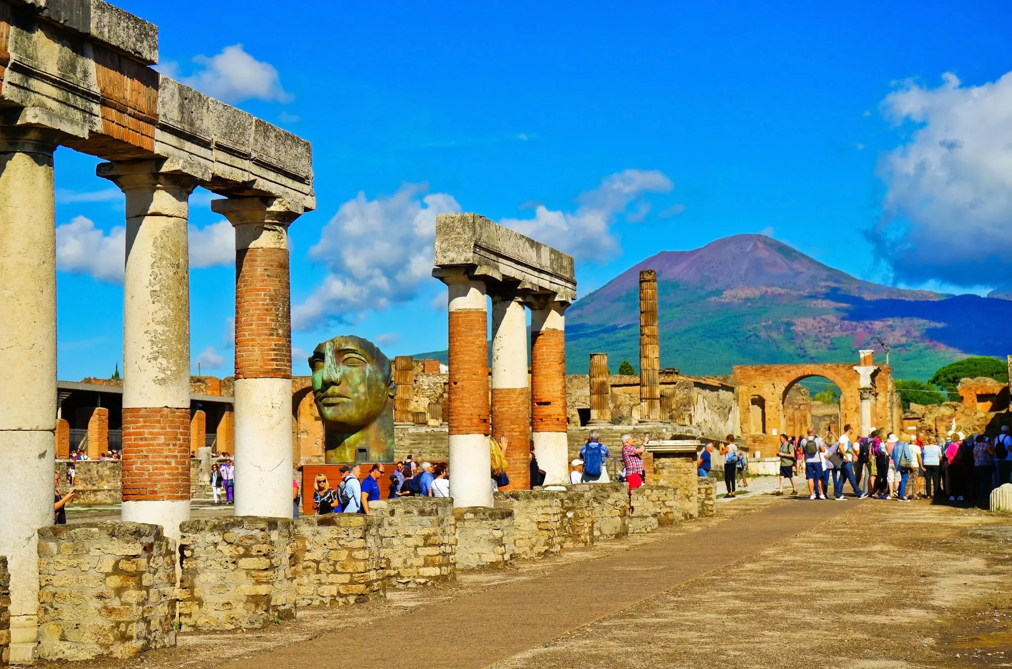 Tourists stand among tall stone columns with a mountain rising in the distance above the town
