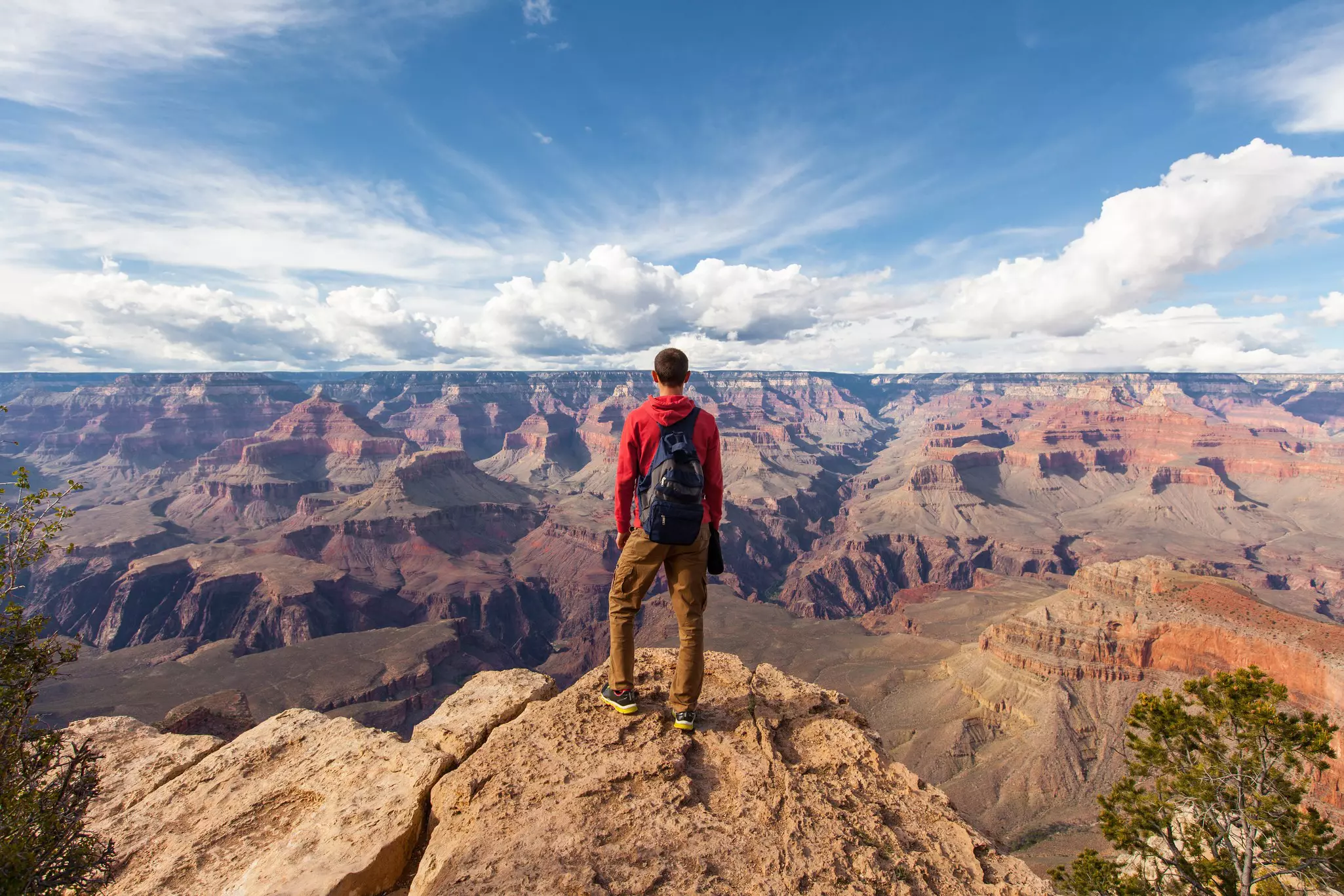 A man in a red sweater and black backpack stands on a rock at the top of a cliff facing a wide view of mesas and rock formations in red, brown and purple.