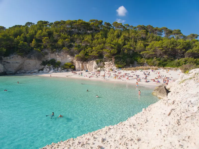 People swim in clear blue waters of a white-sand beach overlooked by white cliffs and trees