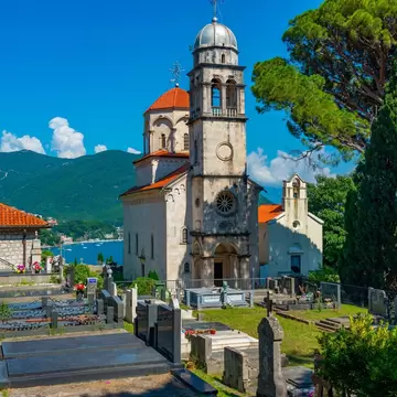 Stone church with a bell tower and red-tiled dome beside a cemetery, pine trees and a blue bay behind.