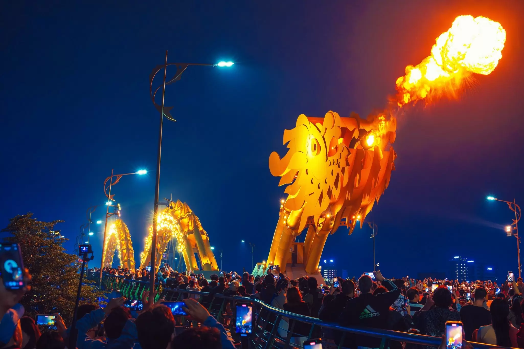 Flames shoot from a bridge structure shaped like the head of a dragon in a city at night as spectators watch, Danang, Vietnam