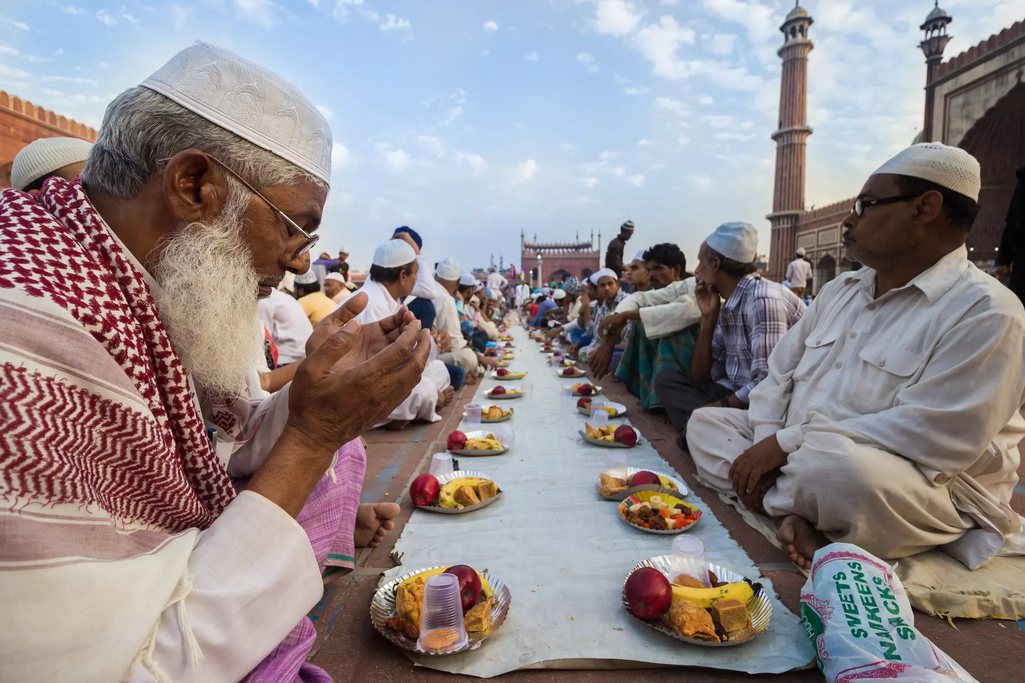 Men in skullcaps sit side by side as they prepare to eat a meal in the courtyard of a mosque.