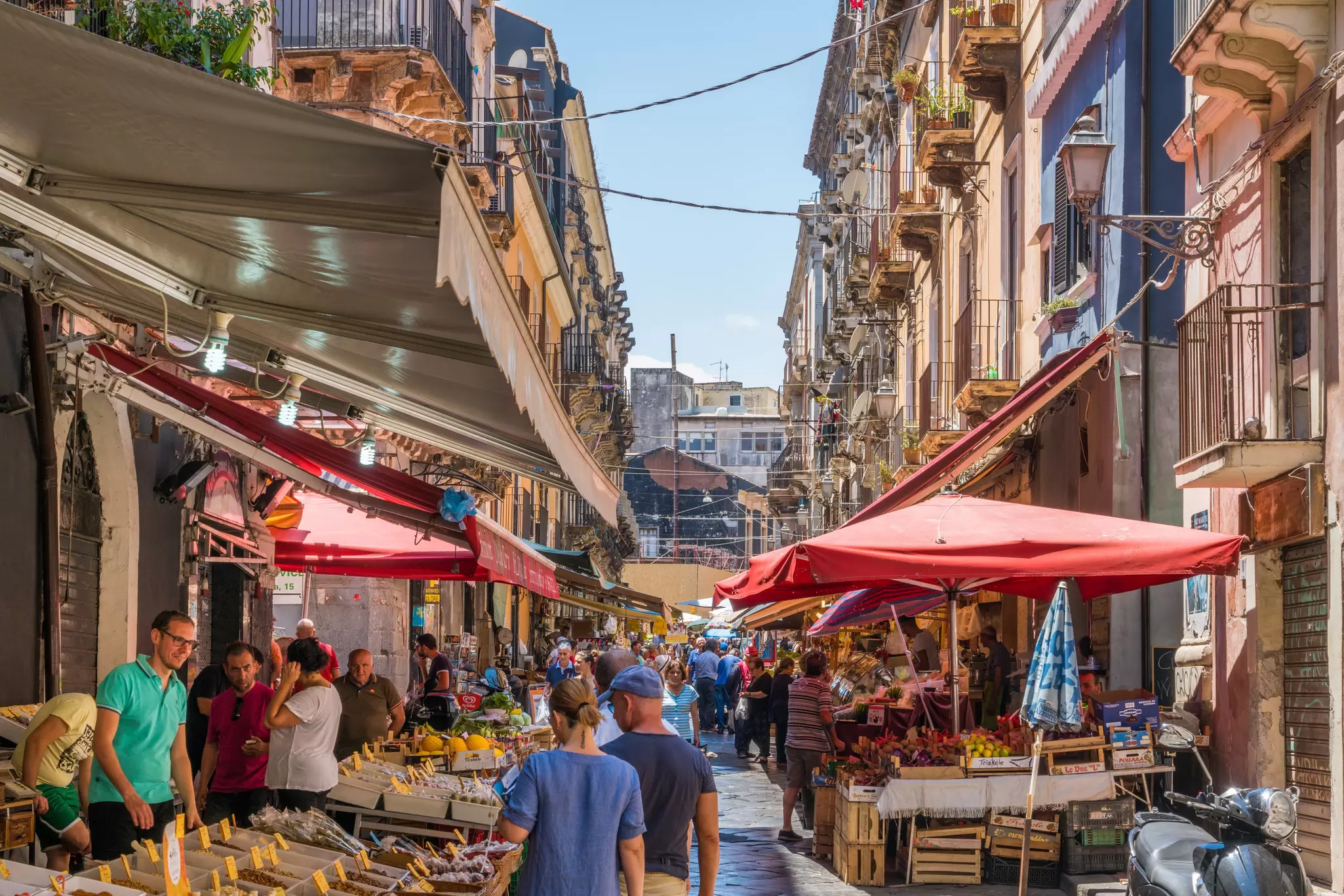 People walk through a street with produce vendors set up on the sidewalk.