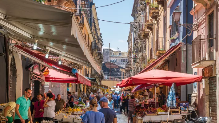 People walk through a narrow street with market stalls on either side; there are red awnings and umbrellas, and displays of produce and other goods on the street.