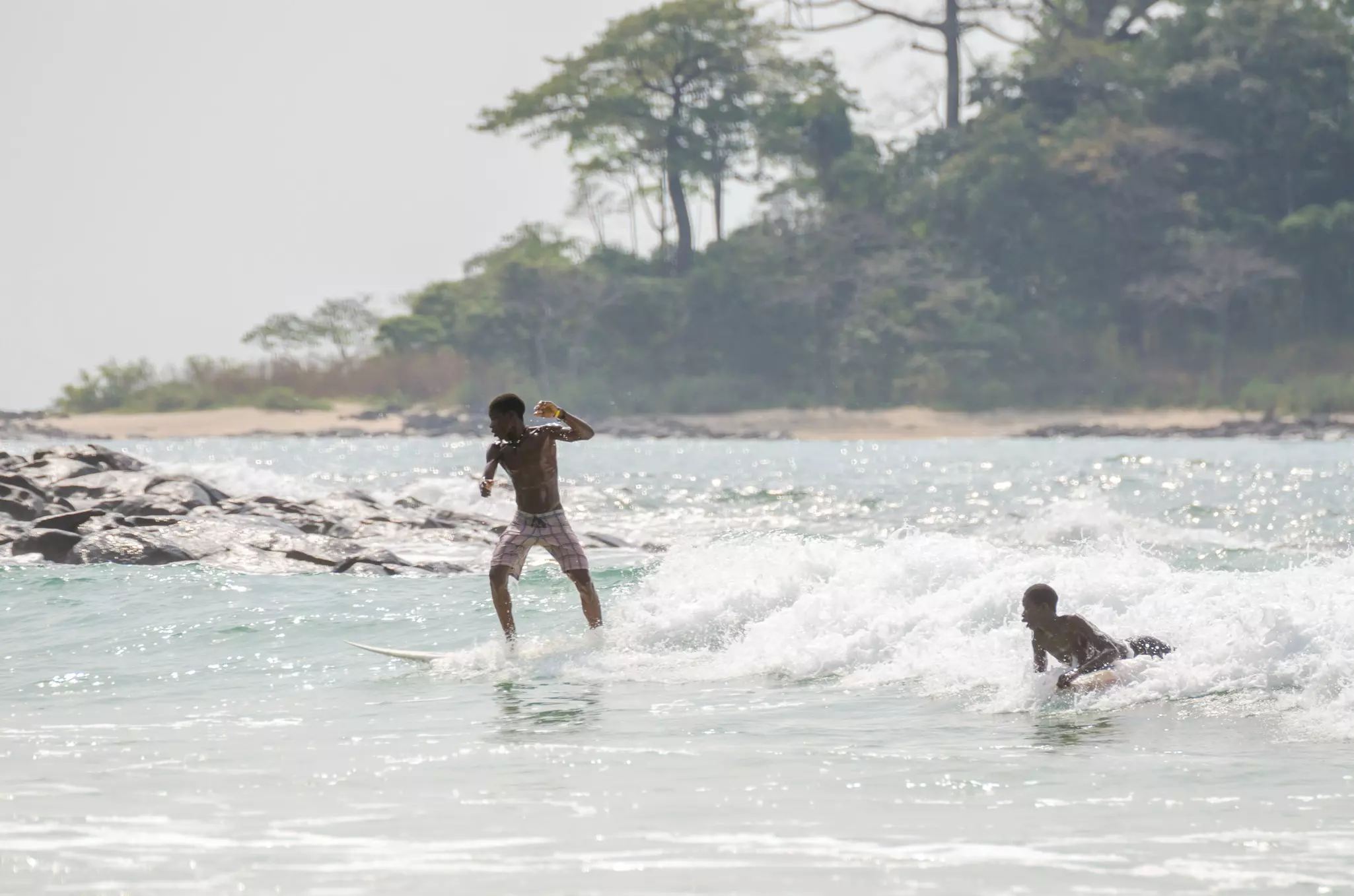 Two boys surf on a small break by a coastline covered in trees.