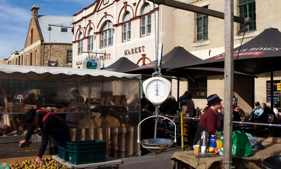 Crates of apples and a hanging scale at an outdoor fruit market stall by historic sandstone buildings.