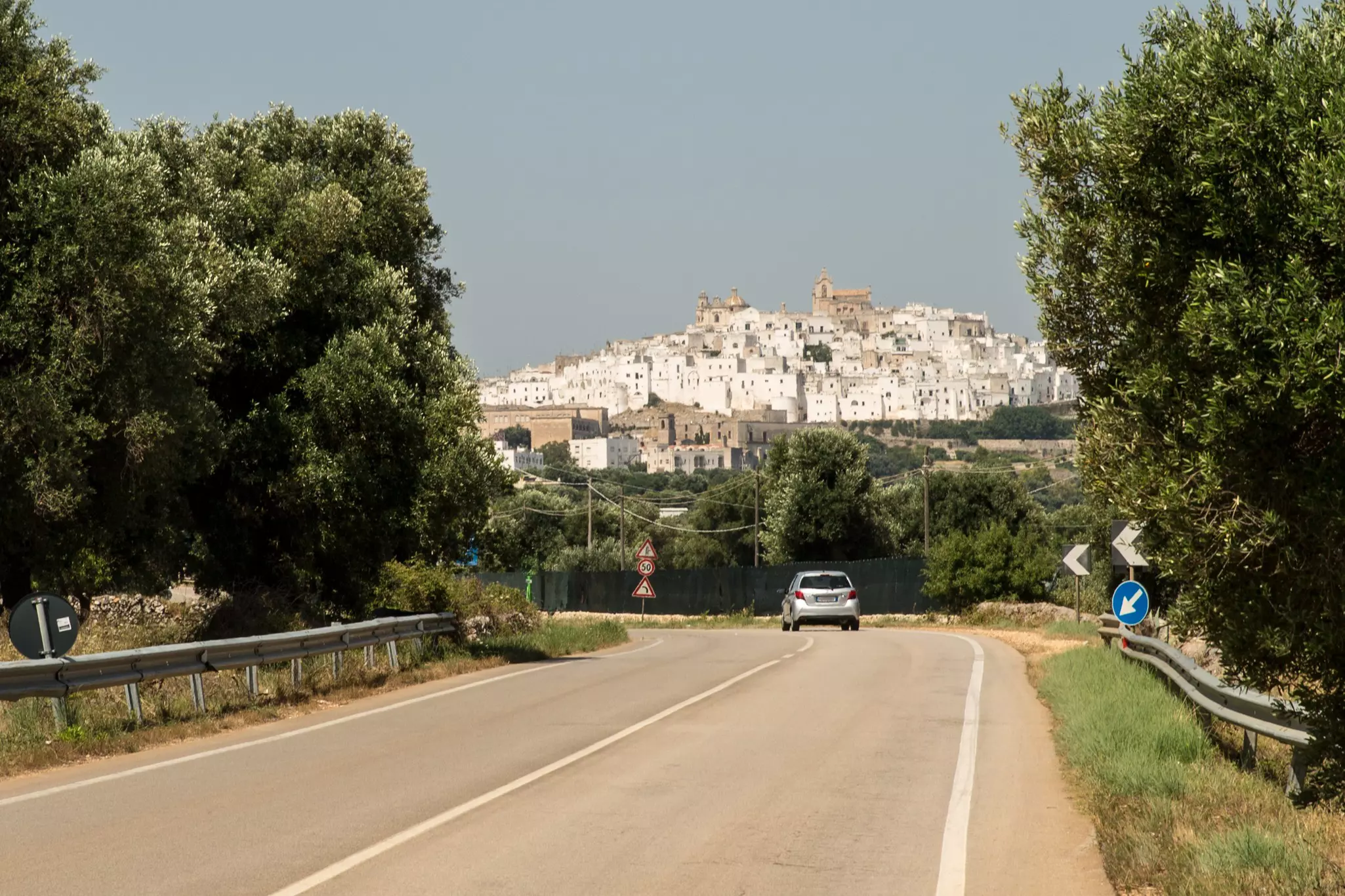 A small car on a road heading towards a hilltop town full of white buildings.