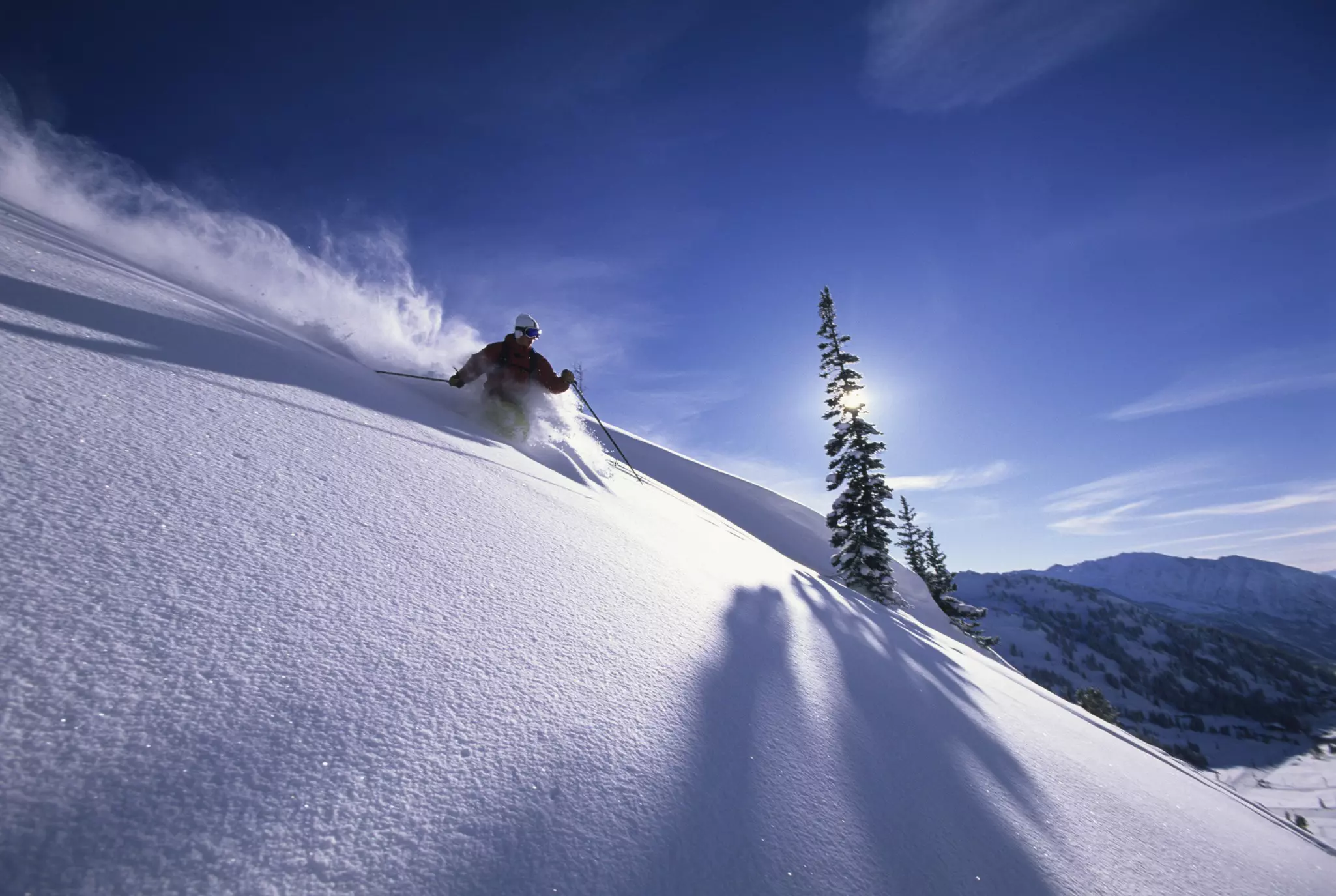 A skier makes a turn on a steep slope, lit by the low rays of the sun.