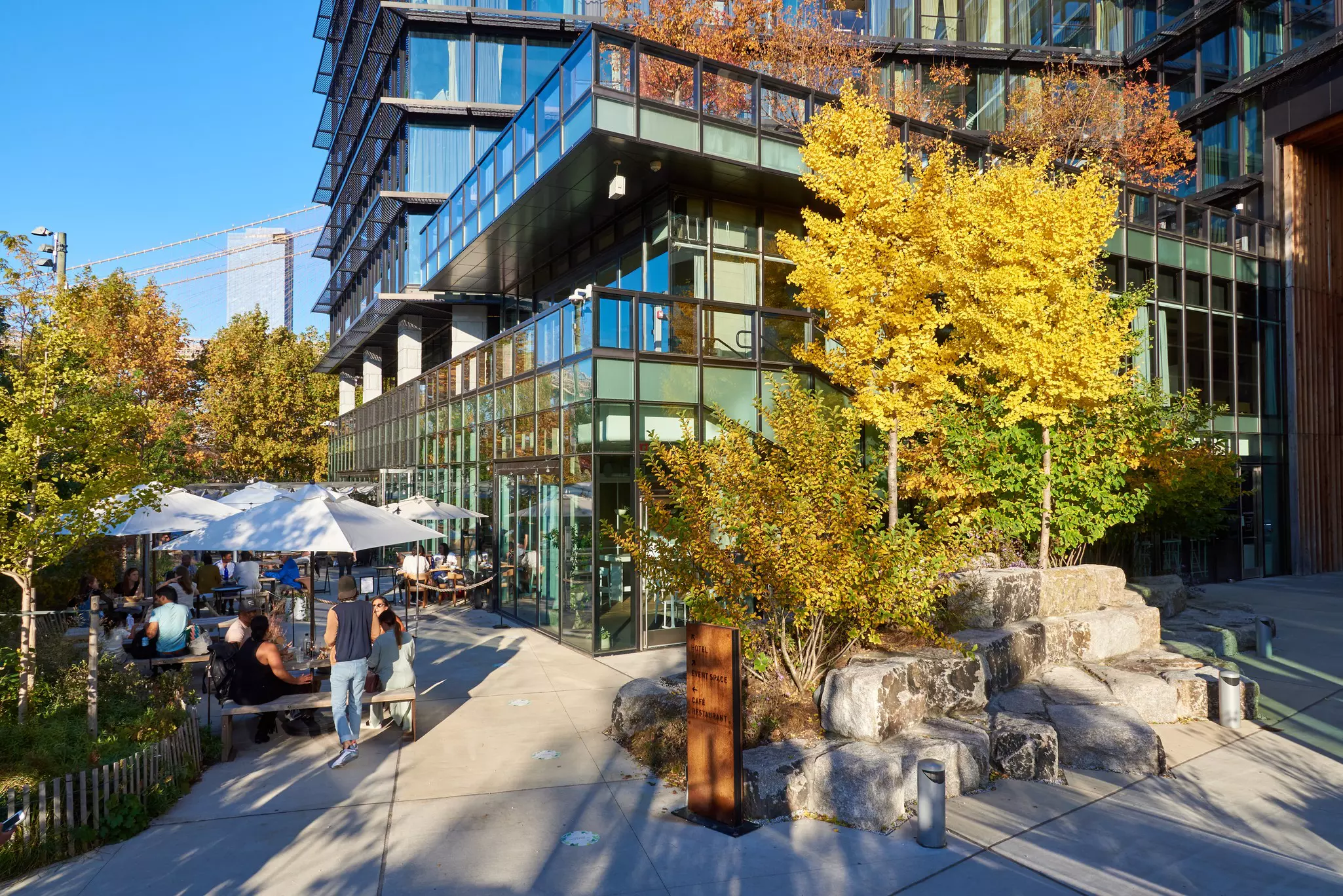 People sit at outdoor tables next to a modern glass hotel set in a city park.