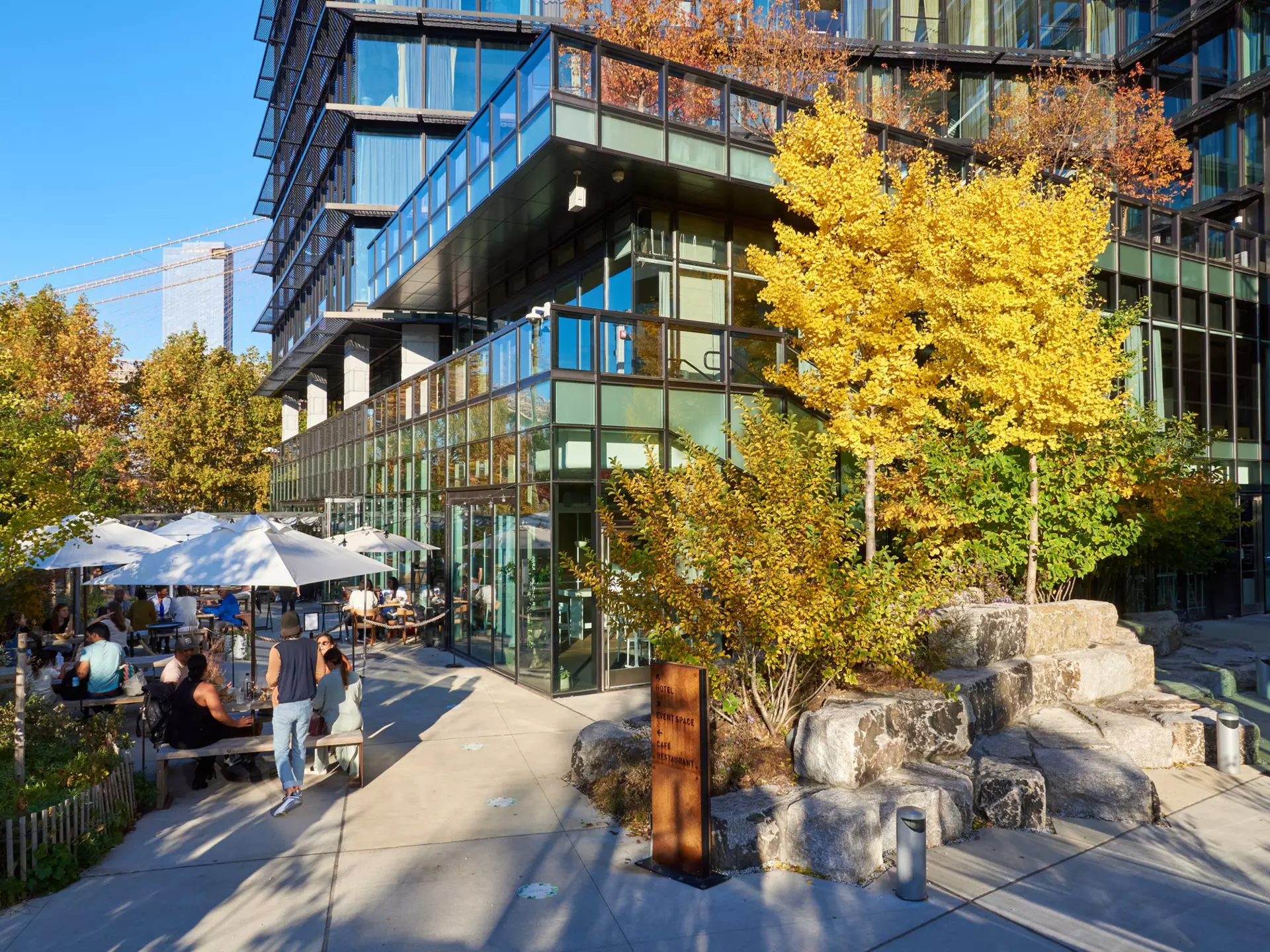 People sit at outdoor tables next to a modern glass hotel set in a city park.