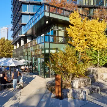 People sit at outdoor tables next to a modern glass hotel set in a city park.