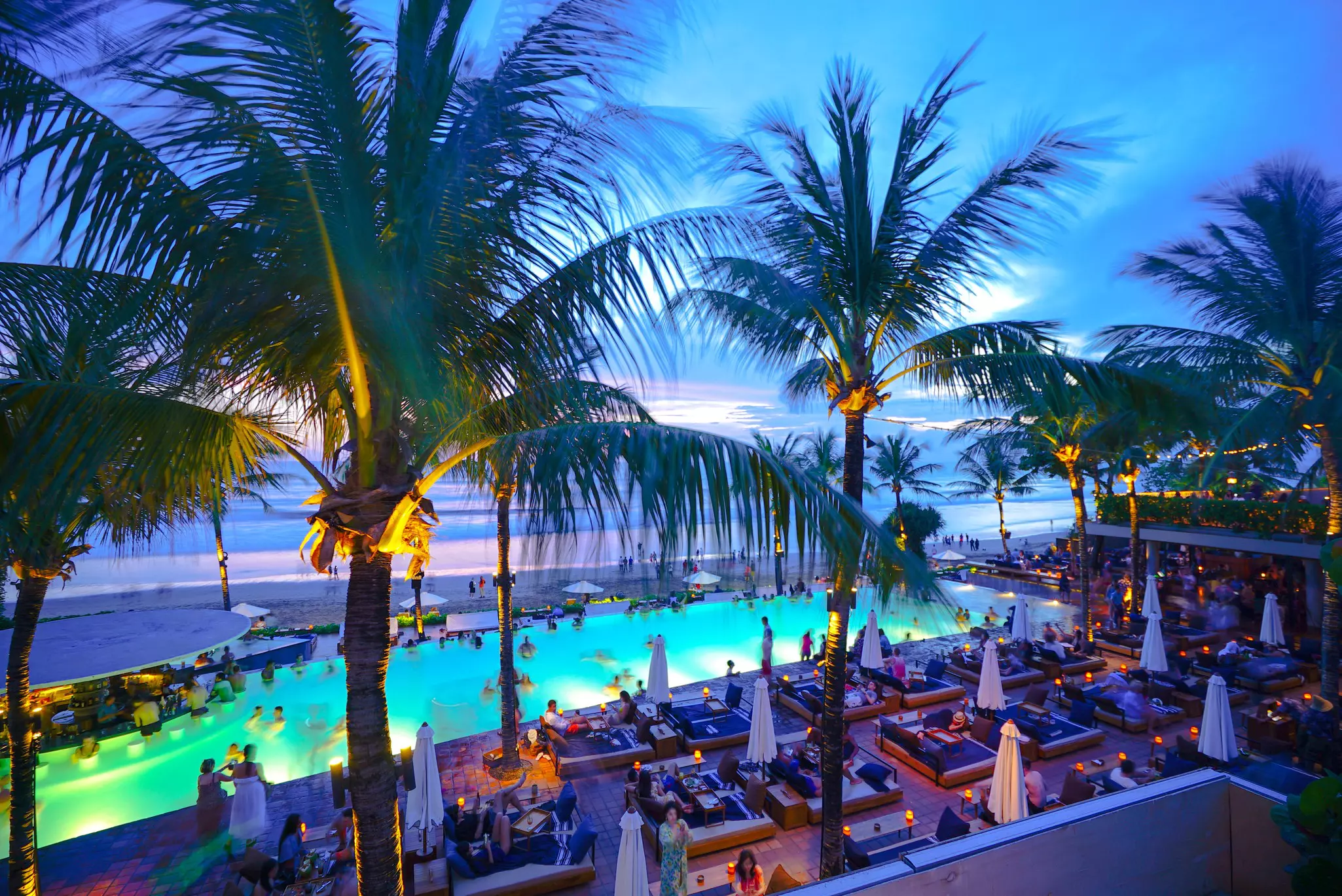 An overhead view of tables surrounding a pool at a club by the beach. Palm trees are in the front of the shot, while the twilight has blue and purple tones.