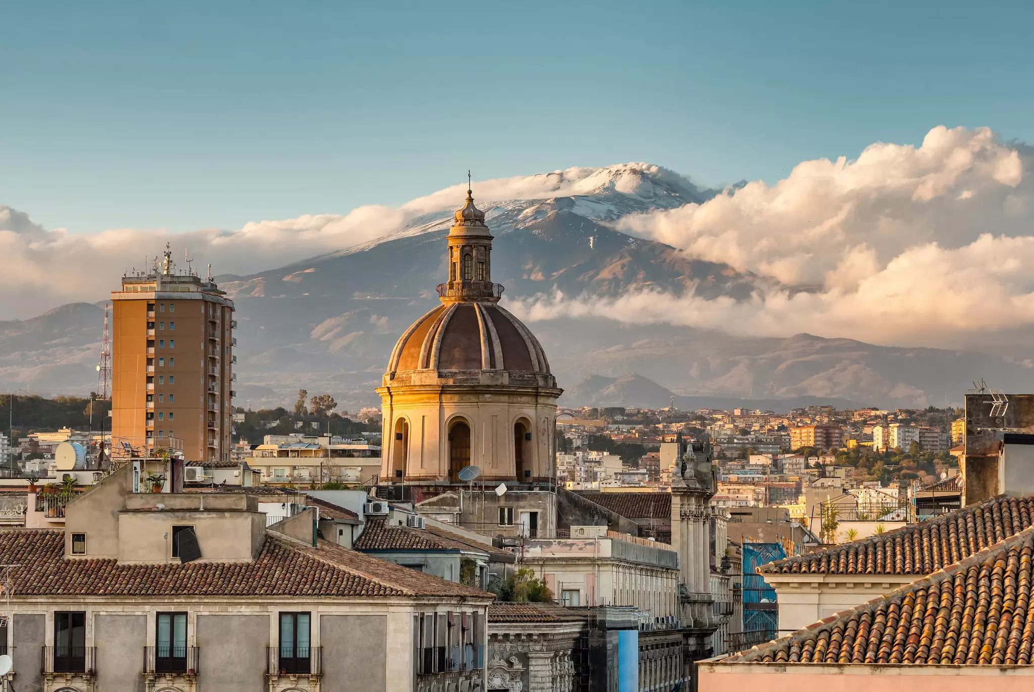 A city glowing in the evening light backed by a snow-capped volcano with clouds rolling off it.