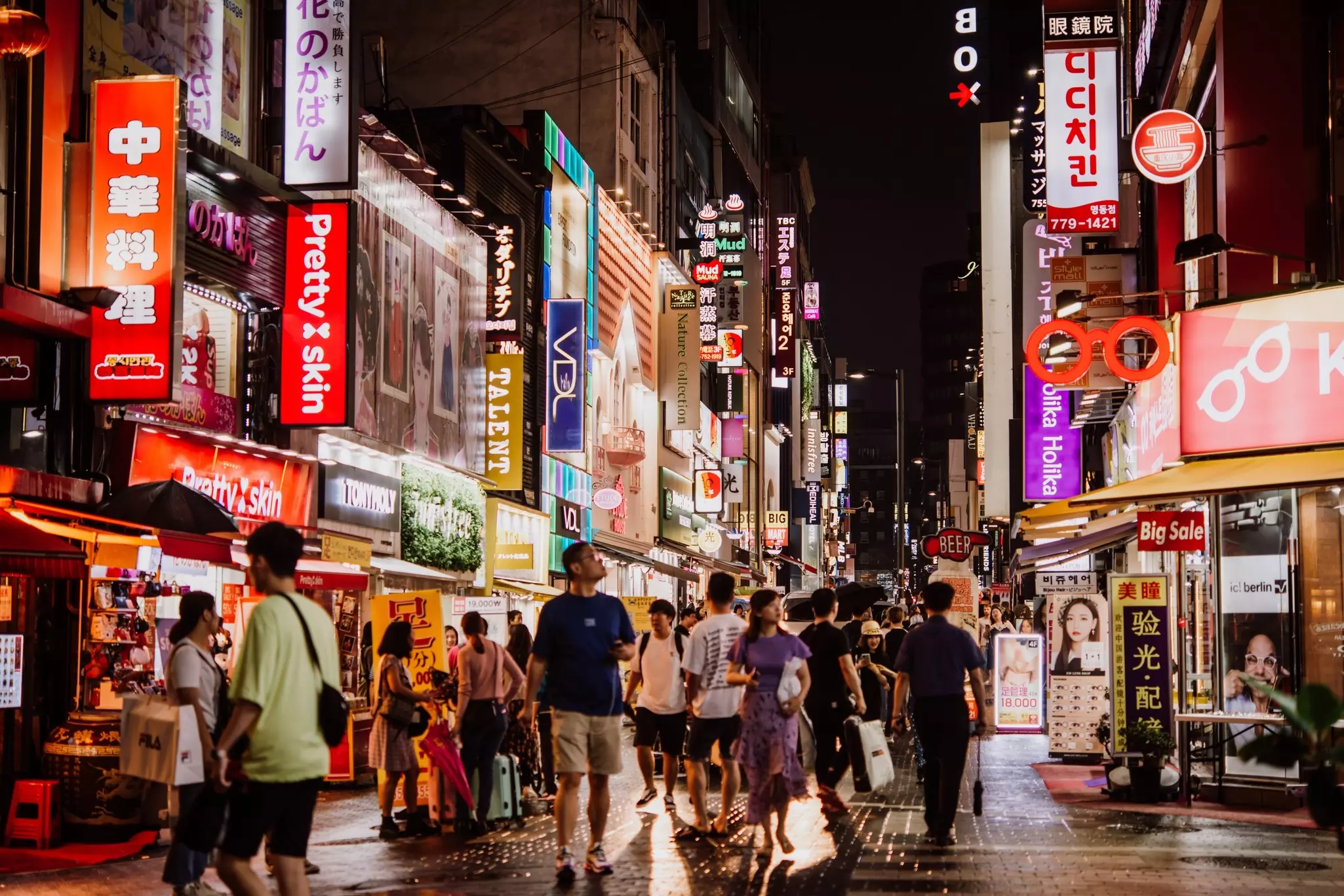 A crowd of people walk on pedestrian-only street at night in a large city. Lit-up signs and billboards flood the street scene with light.