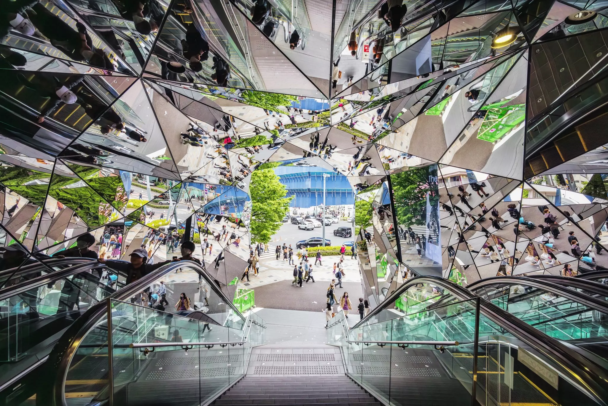 Shoppers and pedestrians reflected in mirrors along an escalator and stairs at a mall entrance