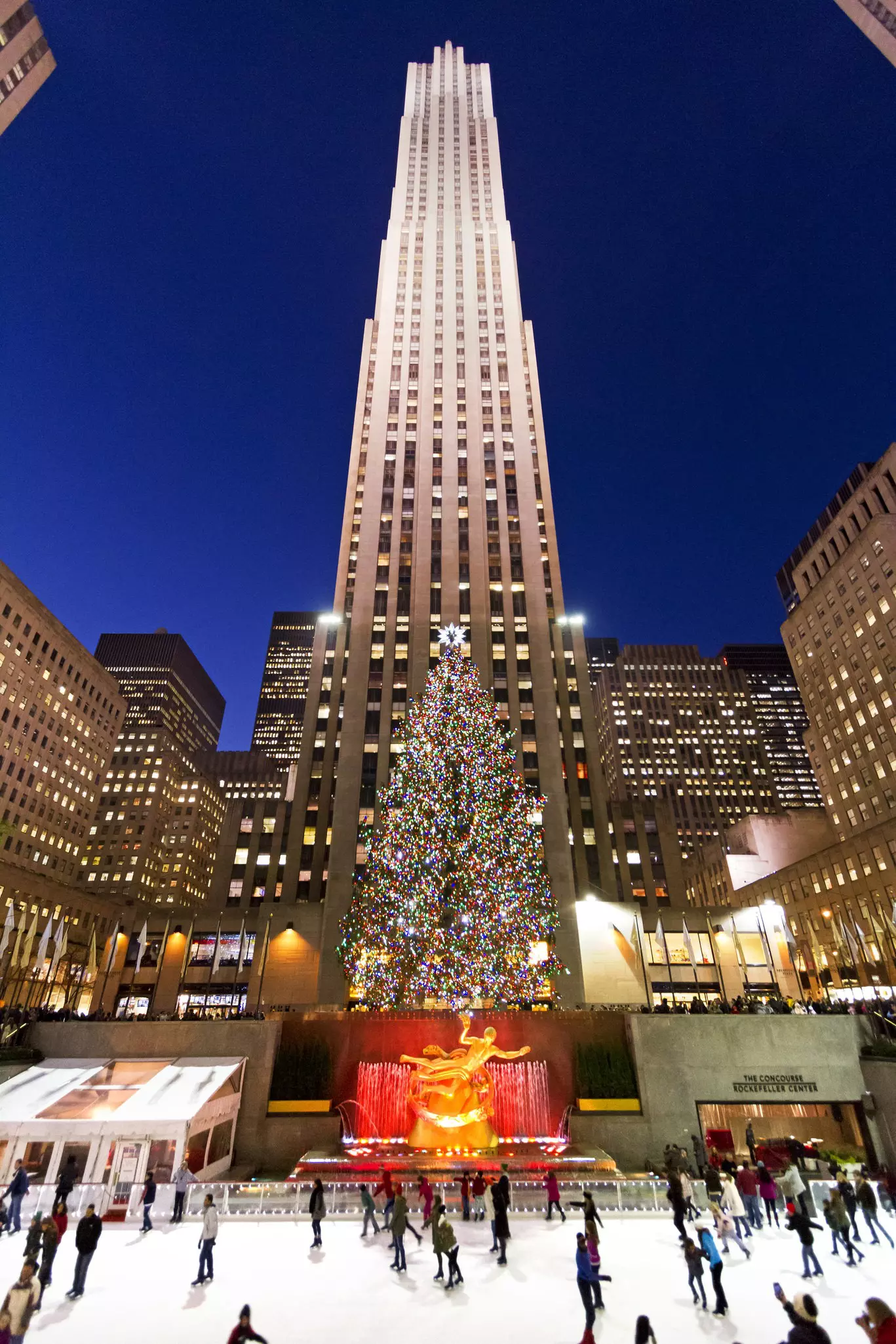 A tall building stands behind a large colorfully lit Christmas tree in front of which is the golden statue of Prometheus while people skate in front