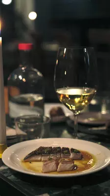 A glass of white wine and a small plate with fish dressed in olive oil are on a candlelit table.