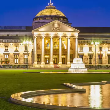 A domed neoclassical building with columns, inscribed Aquis Mattiacis, floodlit at dusk behind a lawn and fountain, Wiesbaden