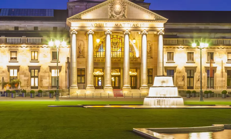 A domed neoclassical building with columns, inscribed Aquis Mattiacis, floodlit at dusk behind a lawn and fountain, Wiesbaden
