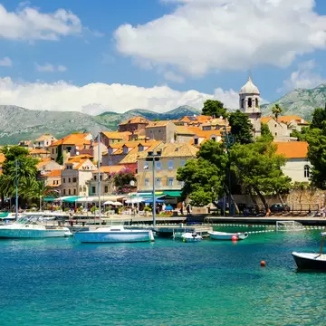 Boats moored in turquoise water in front of orange-roofed houses and a white bell tower in Cavtat, Croatia, mountains behind