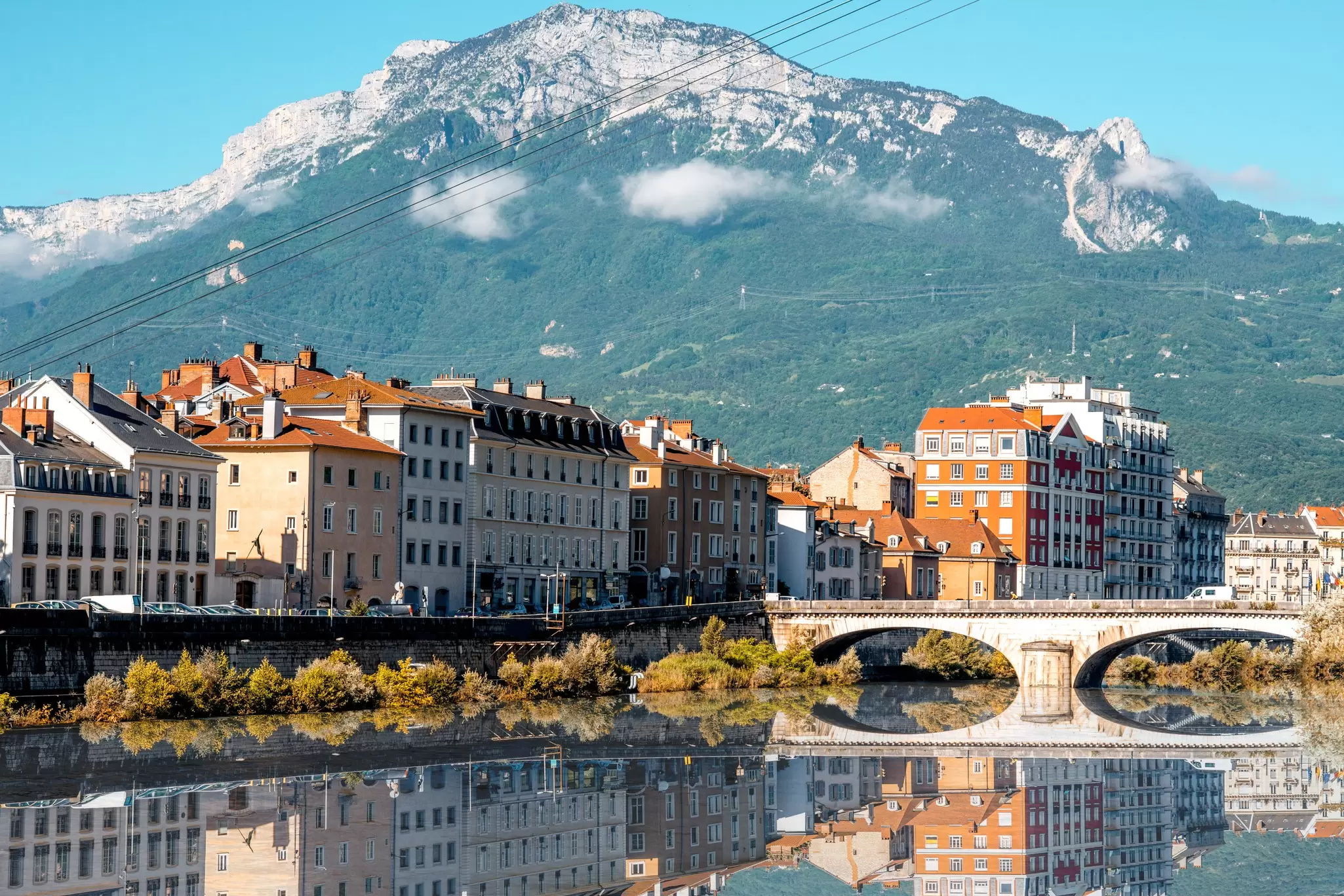 A bridge crosses a river at a town; cable car lines run overhead, and a rocky mountain is in the background.