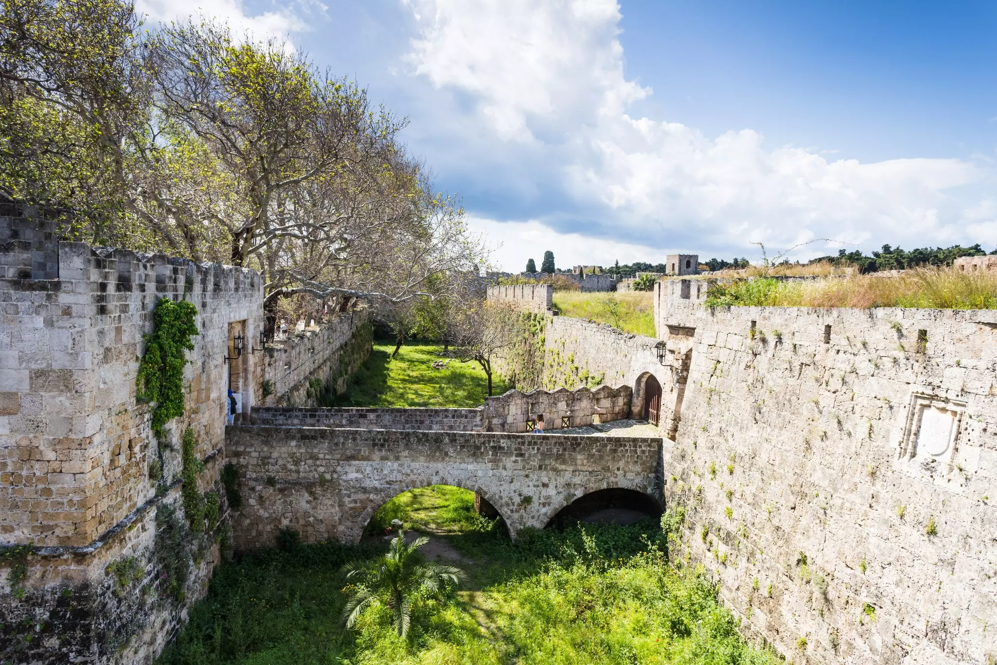 A view of the fortified walls of a medieval city, with a stone bridge crossing a moat now planted with grass and other greenery.