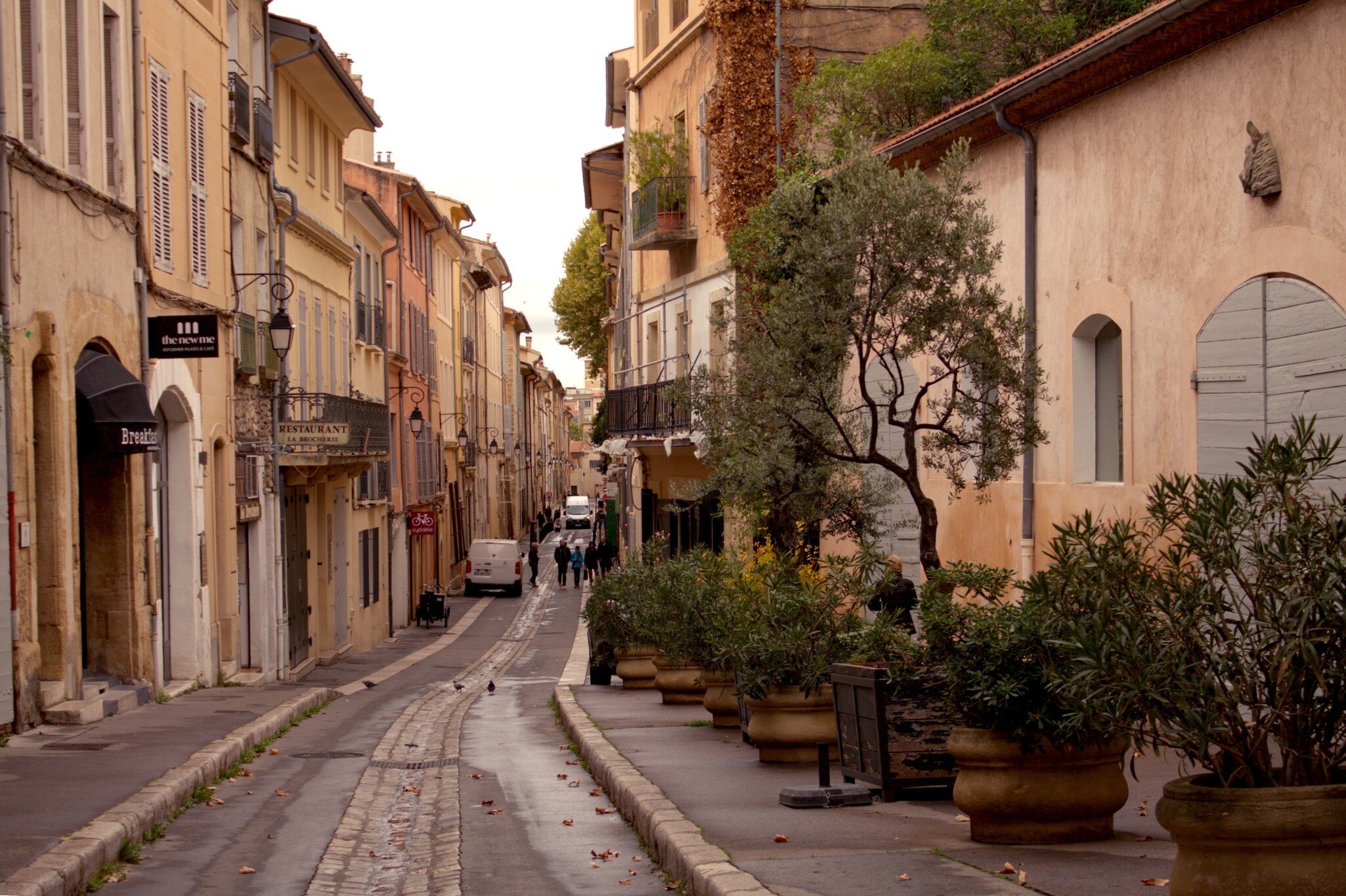 A narrow street of shuttered ocher facades and potted olive trees in Aix-en-Provence; signs read 'Restaurant'