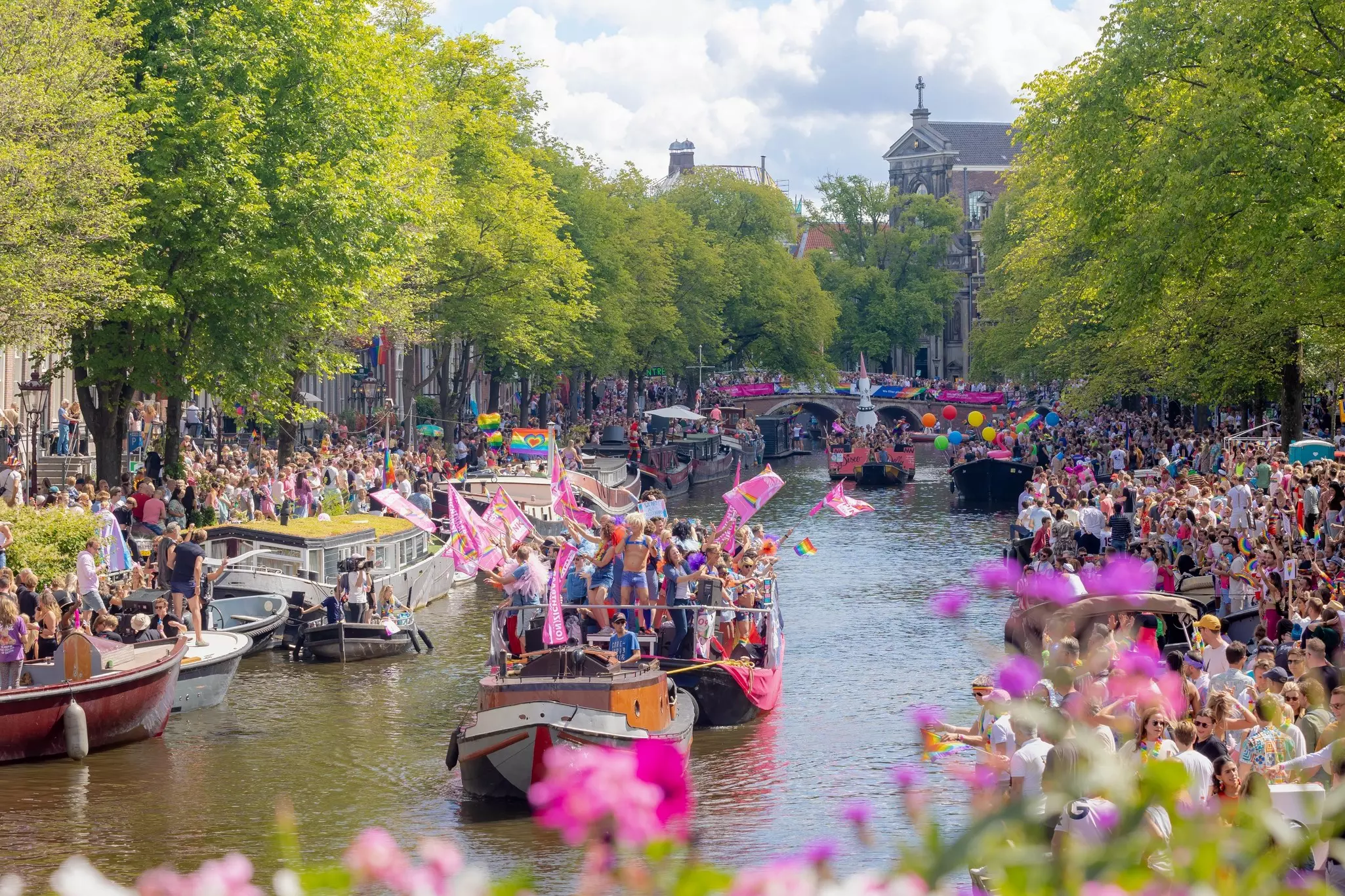 Boats with revelers on decks and lining the canalsides waving rainbow and pink flags during an LGBTIQ+ celebration
