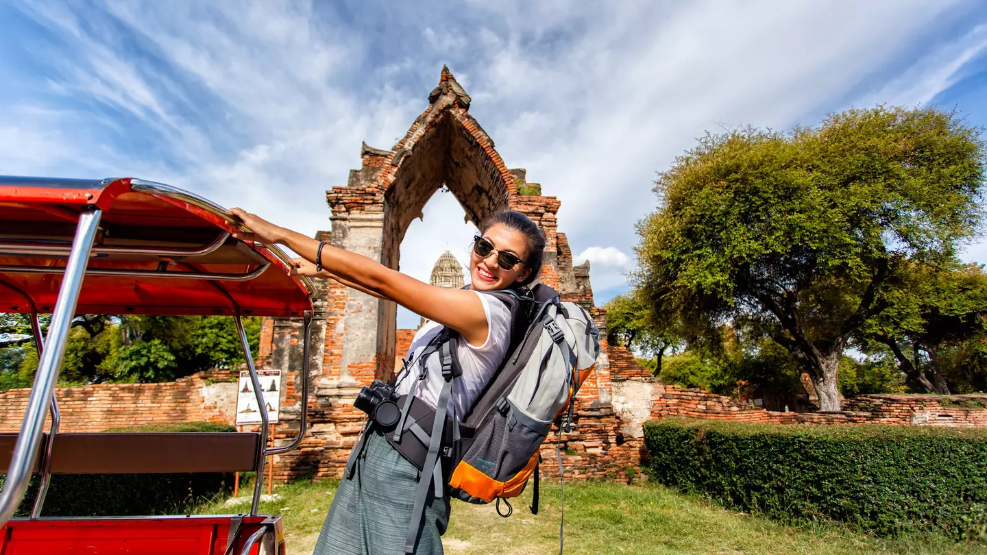 Young asian female traveler with backpack standing on Tuk Tuk with Wat Mahathat in background