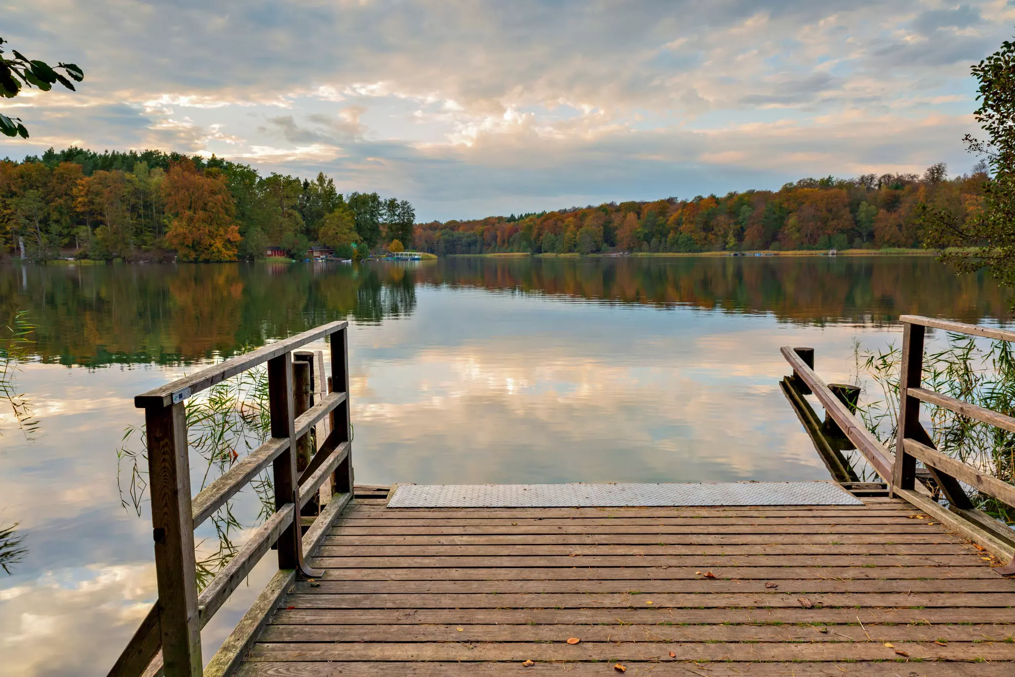 A wooden dock leads to a clear lake that reflects the sky; trees in autumn colors surround the shore.