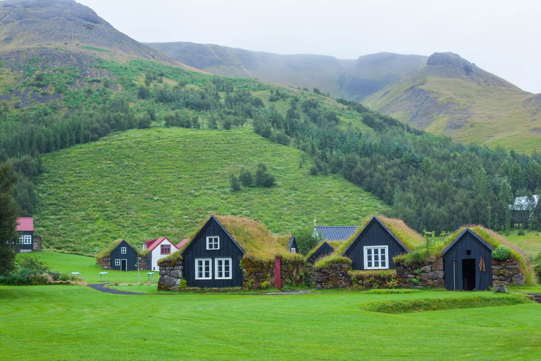 Traditional houses with grass roofs are pictured, against green hills covered in trees and mist.