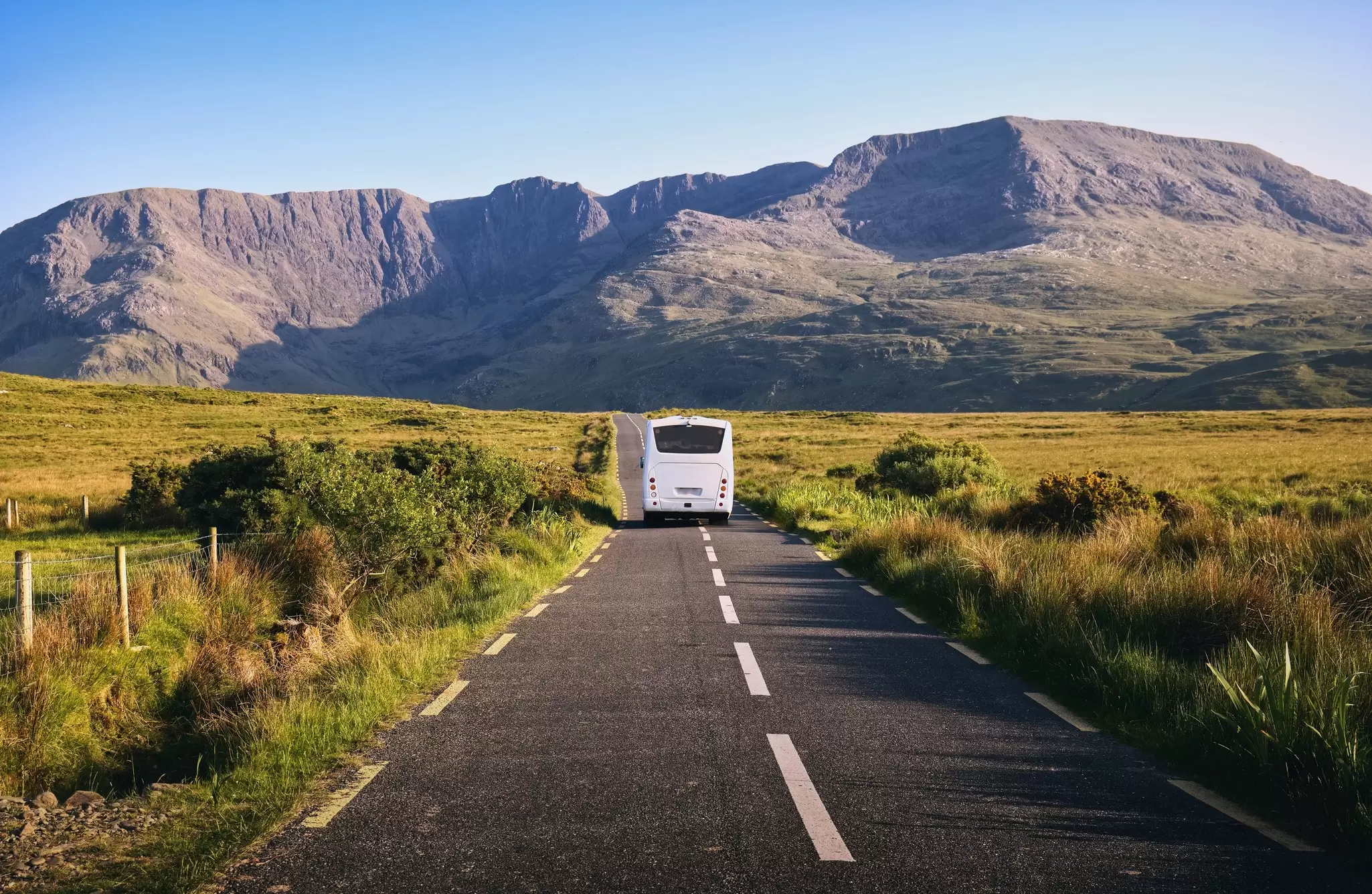 A bus is seen from behind driving on a rural road toward mountains.