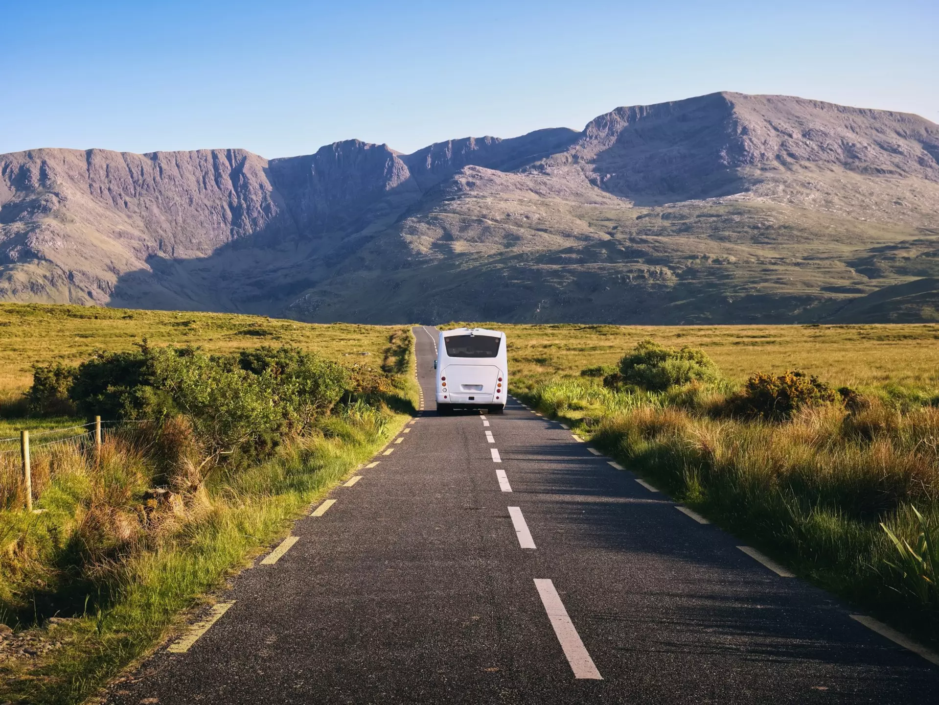 A bus is seen from behind driving on a rural road toward mountains.