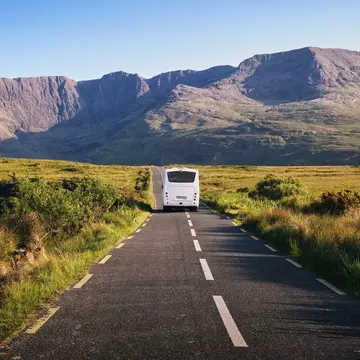 A bus is seen from behind driving on a rural road toward mountains.