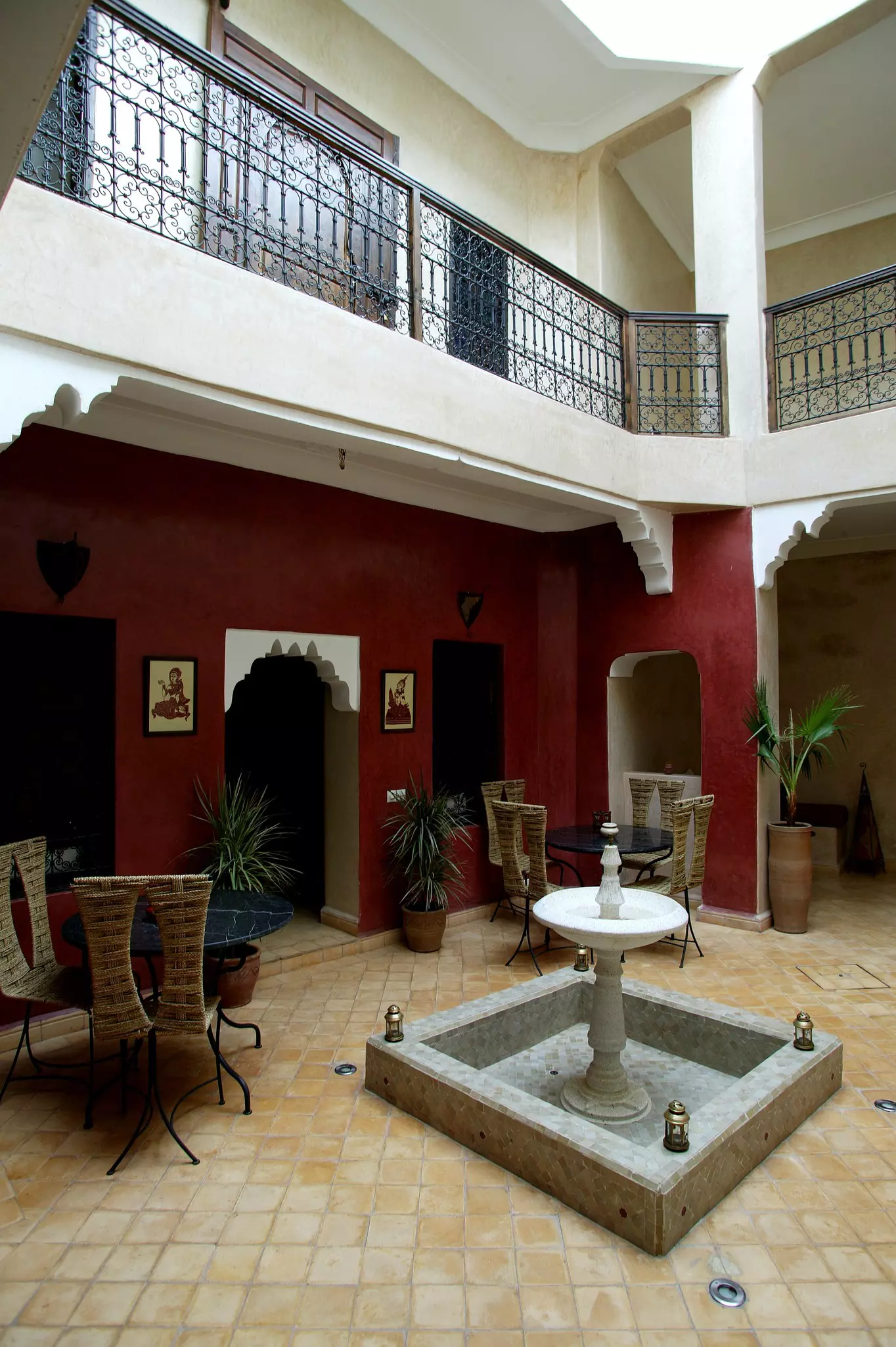 A lobby with beige tiled flooring. There are two round black tables with wicker chairs surrounding them. A square fountain is in the foreground between the tables.