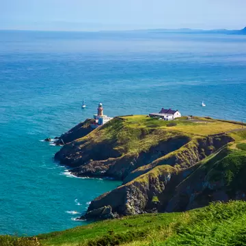 White lighthouse and cottage on a green headland with rocky cliffs, surrounded by blue sea with sailboats.