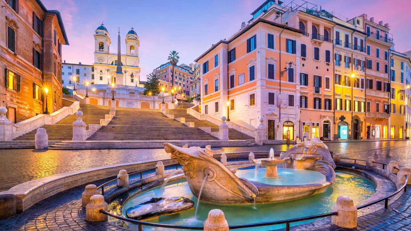 A series of steps lead down to a Roman plaza with cobbled stone streets and in the center of the photo is a marble foundation in the shape of a boat.
